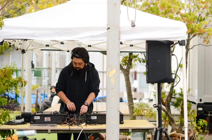 テントの下で機材を操作するDJと、その奥に広がる会場