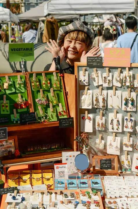 手づくりのイヤリングがずらりと並ぶブースと、笑顔でポーズをとる出店者